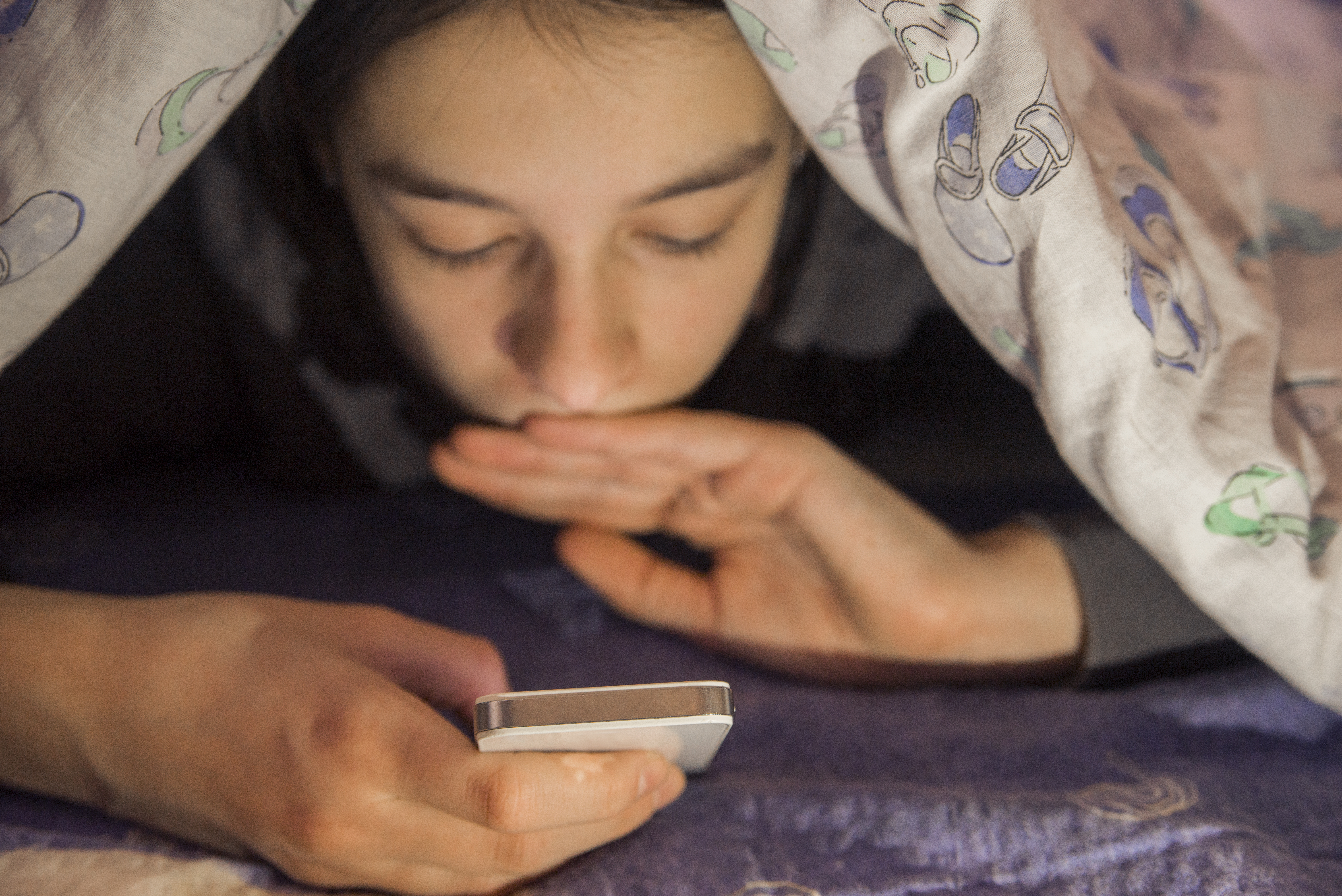 Jente ligger under dynen og leser på mobiltelefon.