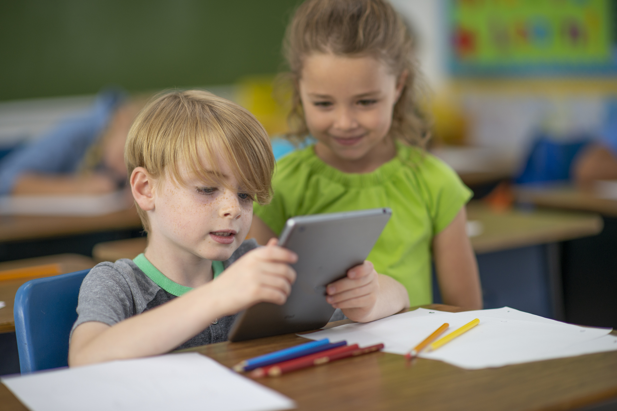 Gutt og jente med ipad i klasserommet. Gutten holder ipaden. Jenta står ved siden av og ser på.