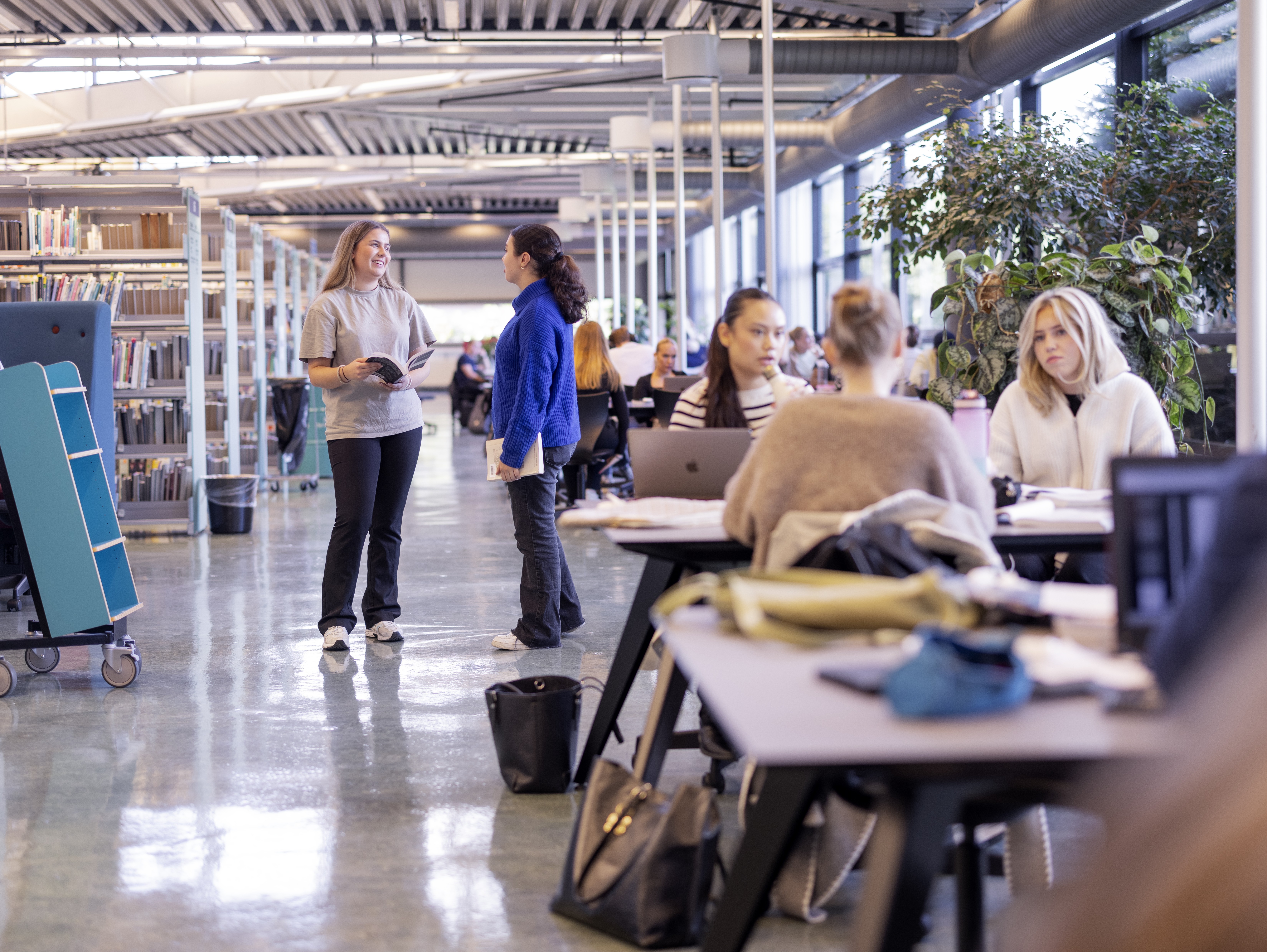 noen elever studerer ved bord, mens to elever står og snakkes sammen