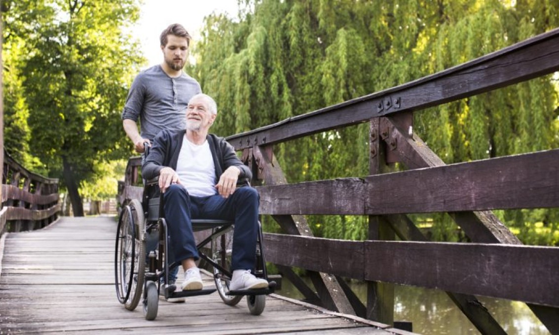 Sønn triller far i rullestol over en bro i en park