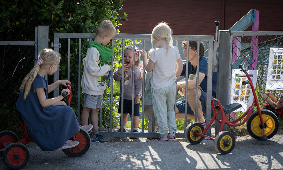 Gråtende jente på utsiden av en port sammen med en voksen. Tre nysgjerrige barn innenfor poten