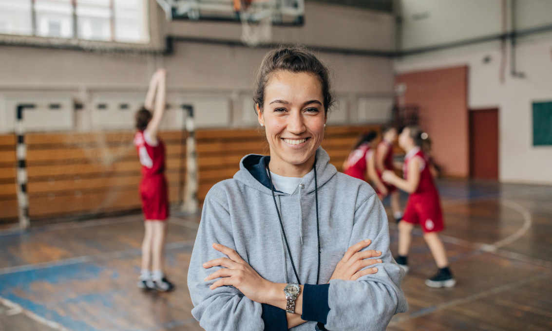 ung kvinne med med håret i hestehale står i en gymsal, smiler til kamera. Tre barn holder på med ball i bakgrunnen