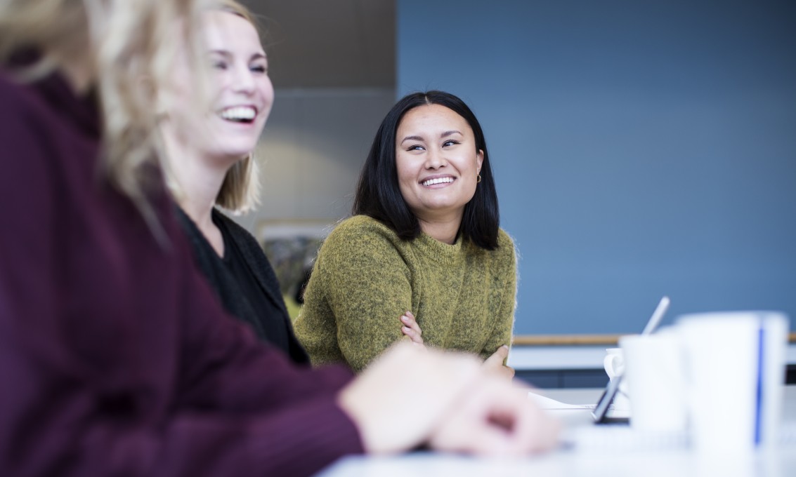 Tre kvinnelige studenter sitter på rekke og ser smilende på hverandre.