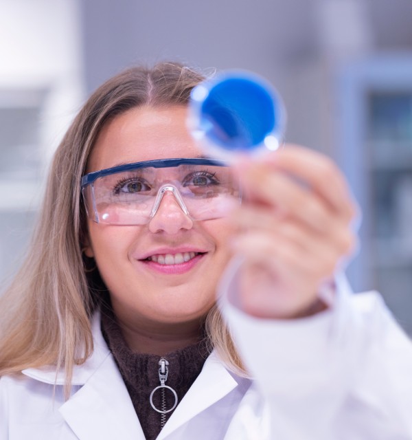 Kvinnelig student iført lab frakk og vernebriller som ser på en beholder med noe blått i