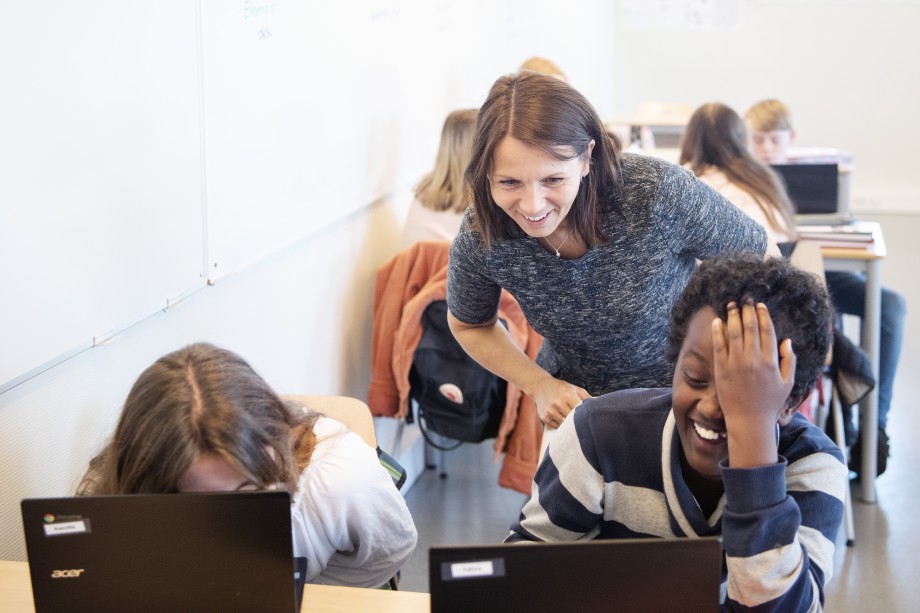 Smilende lærer ser over skulderne på to elever som ler av noe de ser på pc-ene sine.