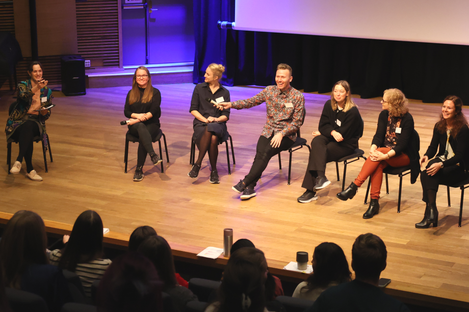 Fotografi av syv smilende personer som sitter på hver sin stol på en scene i forbindelse med en paneldebatt.