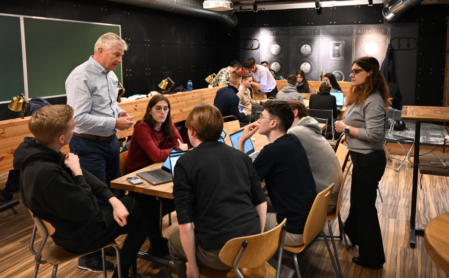 En mann i skjorte forklarer noe til en gruppe studenter rundt et bord.