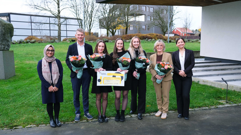 Sju personer står ved bygg med blomster og gavesjekk