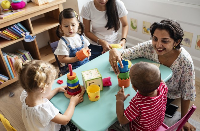 Tre små barn sitter rundt et bord med en voksen mens de leker med klosser